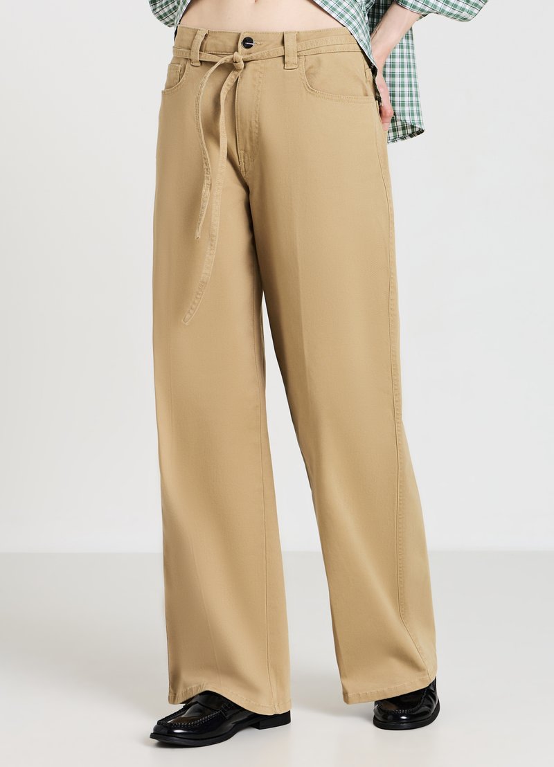 Pantalon beige à jambes larges avec taille à cordon, poches latérales et texture lisse, associé à des mocassins noirs.