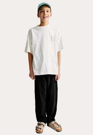 T-shirt en coton blanc avec détail de poche, pantalon noir ample et sandales beiges à double boucle, complétés par une casquette bleu clair.