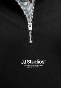 Czarna bluza z suwakem, z metalowym zamkiem błyskawicznym, z białym napisem "JJ Studios" oraz paskowanym kołnierzem. Miękka dzianina.