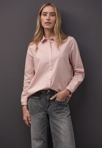 Světle růžová košile na knoflíky s tenkými vertikálními pruhy, kombinovaná s tmavě šedými džínami. Košile má límec a dlouhé rukávy.