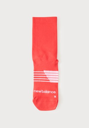 Hellrote New Balance Socke mit weißen diagonalen Streifen und Logo-Text nahe dem Zehenbereich auf hellgrauem Hintergrund.