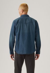 Denim shirt in deep blue with long sleeves, button cuffs, and a curved hem. Features a back yoke and subtle variations in shade.