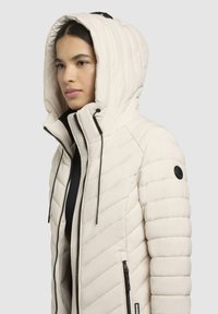 Letvægts, cremefarvet pufferjakke med quiltet design, høj krave og justerbar hætte; har sort hardware og lynlåse.