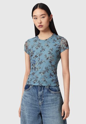 Ung kvinde med glat sort hår iført en blå blomstret kortærmet top og højtaljede løse denimjeans mod en lys baggrund.