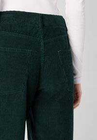 Grüne Cordhosen mit gerippter Textur, mittelhoher Taille und zwei Gesäßtaschen, aus einem hinteren Winkel aufgenommen, mit einem sichtbaren weißen Oberteil.