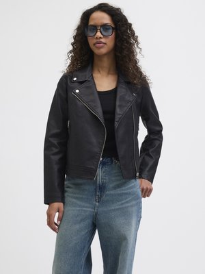 Vrouw met krullend haar die een zwarte zonnebril draagt, een zwart leren jack, een zwart topje en hooggesneden blauwe jeans, staand tegen een effen achtergrond.