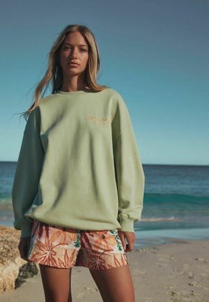 Noor naine seisab liivarannal, kandes helesinist pusa ja lillemustriga lühikesi pükse, taustal meri ja selge taevas.