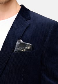 Marineblå kordfløyelsblazer med knapper på kragen, som har en svart lommetørkle med sommerfugl-mønster liggende i brystlommen.