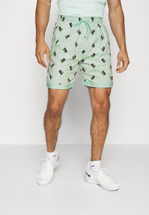 Mann trägt hellgrüne Shorts mit Kordelzug und kleinen schwarzen Hundedrucken, kombiniert mit weißen Nike-Sneakers und weißen Knöchelsocken.