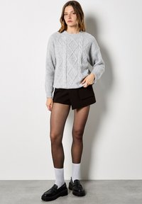 Szary sweter dziany z wzorem w warkocze, zestawiony z czarnymi szortami i czarnymi masywnymi mokasynami. Modelka nosi półprzezroczyste czarne rajstopy oraz białe skarpetki.