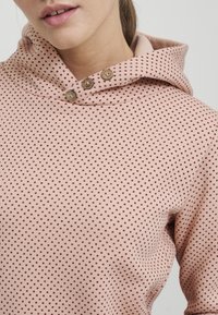 Sudadera de color rosa claro con un patrón de puntos en rojo oscuro. Cuenta con una gran capucha y tres botones decorativos en el cuello. Textura de tela suave.
