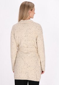 Cardigan beige con un motivo testurizzato, caratterizzato da macchie nere, maniche lunghe, vita con cintura e collo scialle. Tessuto lavorato a maglia morbido.