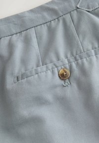 Közeli kép egy világosszürke szövet nadrág zsebének részletéről, melynek a derékszíj alatti hurkon egy barna gomb van rögzítve.