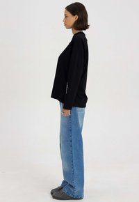 Czarna koszula z długim rękawem o luźnym kroju, z logo po boku, połączona z jasnoniebieskimi spodniami typu wide-leg i ciemnymi sneakersami.