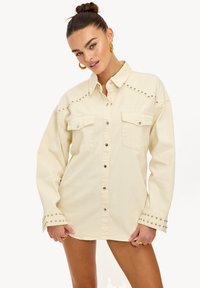 Vestido camisero de denim color beige claro con acentos de tachuelas en los hombros y puños, con dos bolsillos frontales y diseño de botones.