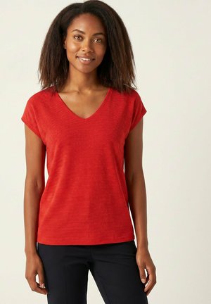 Femme aux cheveux naturels souriante, portant un haut rouge à manches courtes avec col en V et un pantalon noir, debout sur un fond clair uni.