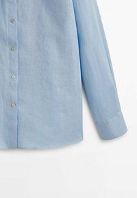 Chemise bleu clair à manches longues avec boutons et manchettes retroussées, présentée partiellement sur un fond blanc.