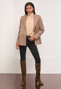 Dvojřadový hnědý houndstooth blazer přes béžový rolák, v kombinaci s černými skinny džíny a hnědými semišovými botami do kolen.