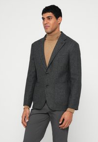 Blazer cinzento escuro de mistura de lã com lapela entalhada, fecho de dois botões, dois bolsos frontais e trama texturizada, usado sobre um gola alta castanha clara.