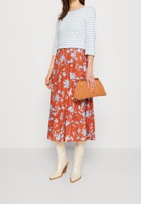 Gestreiftes hellblaues Langarmoberteil kombiniert mit einem roten, floralen Midirock, beigefarbenen kniehohen Stiefeln und einer braunen Clutch.