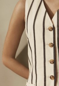 Gilet en lin crème avec des rayures verticales noires, présentant un décolleté en V, cinq boutons et des poches latérales. Texture lisse avec des accents naturels.