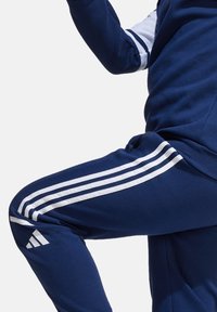 Marineblauwe trainingspak met witte strepen op de mouwen en benen; heeft het Adidas-logo in het wit op de dij van de broek. Zachte, sportieve uitstraling.