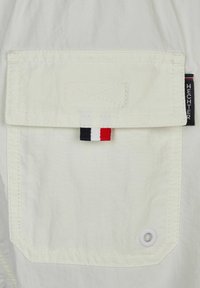 Witte stof met een grote zak en gestikte details; heeft een meerkleurig label (rood, wit en blauw) en een witte ring.