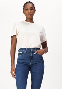 Kreminės spalvos medvilnės marškinėliai su subtiliu "Calvin Klein" logotipo įvaizdžiu; derinami su tamsiai mėlynomis aukšto juosmens džinsinėmis kelnėmis, turinčiomis priekinėmis kišenėmis ir metaline knopke.