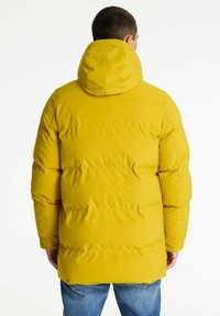 Gul polstret jakke med hætte, med en quiltet design og lodrette sømme, parret med denim jeans.