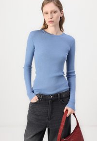 Light blue ribbed long-sleeve top paired with dark gray high-waisted jeans, held with a red bag. Smooth texture and fitted design.