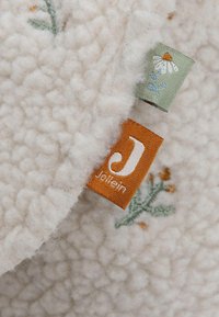 Witte textuurstof met een pluizige afwerking, versierd met geborduurde bloemmotieven. Bevat twee stoffen labels, één oranje met "J" en één groen.