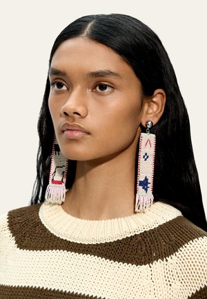 Młoda kobieta z długimi czarnymi włosami nosi duże kolczyki z koralików z geometrycznymi wzorami oraz brązowo-kremowy sweter w paski.