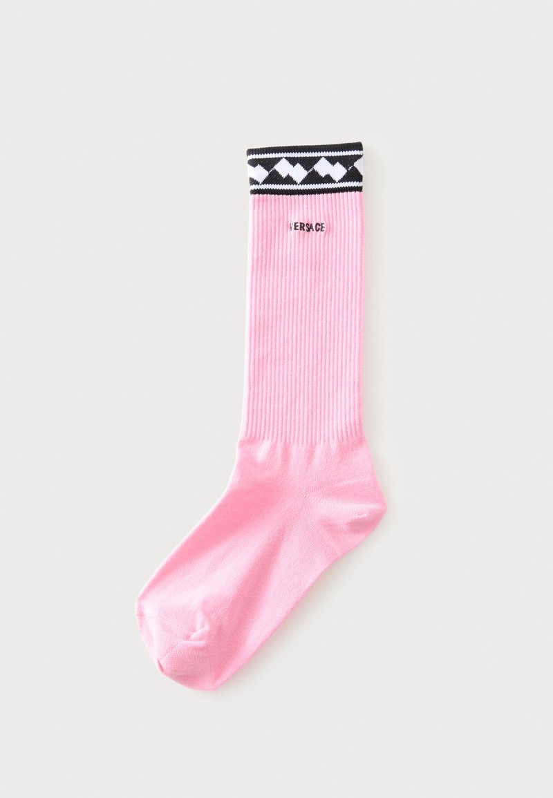 Chaussette côtelée rose avec bord à motif géométrique noir et blanc et logo "Versace" partiellement visible près du haut.