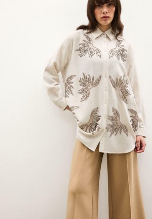 Femme portant une chemise beige à boutons avec des motifs de feuilles marron et un pantalon large couleur sable, se tenant devant un fond clair uni.