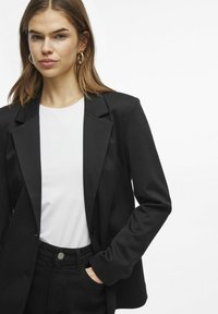 Černý blazer se strukturálním střihem, klopovými límci, zapínáním na jeden knoflík a hladkou texturou. Nositý přes bílou košili a černé kalhoty.