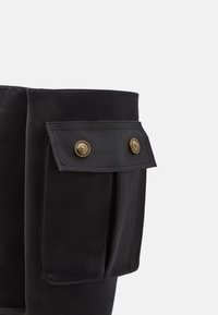 Gros plan sur la tige d'une botte noire présentant une grande poche à rabat rectangulaire avec deux boutons décoratifs dorés.