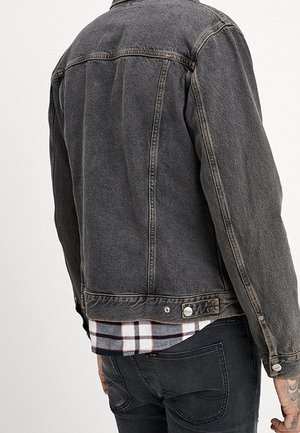 Mężczyzna w wyblakłej czarnej kurtce dżinsowej na białej i czarnej koszuli w kratę oraz czarnych jeansach, pokazany od tyłu i z prawej strony.