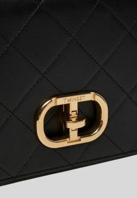 Sac à main en cuir matelassé noir avec un fermoir logo en ton doré au design ovale. Le matériau a une texture lisse et des coutures détaillées.