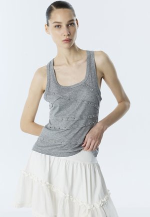 Jonge vrouw die een grijs mouwloos topje met studs en een witte gelaagde rok draagt, staand met één hand op haar heup tegen een lichte achtergrond.