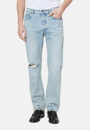 Osoba w jasnoniebieskich podartych dżinsach i czarnych butach, z jedną ręką w kieszeni, a drugą opuszczoną wzdłuż ciała.