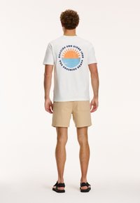 Tricou alb din bumbac cu un grafic al unui soare, raze în degrade de portocalie și albastră; asociat cu pantaloni scurți bej. Design simplu cu o croială lejeră.