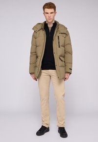 Chaqueta de plumas color caqui con cierre frontal, dos bolsillos laterales, capucha desmontable y acabado texturizado. Combinada con pantalones beige y zapatillas negras.