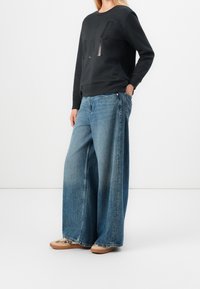 Nainen pukeutuneena mustaan pitkähihaiseen paitaan, jossa on hillitty logo, leveälahkeisiin sinisiin farkkuihin ja beigen värisiin tennareihin, joissa on ruskeat pohjat, seisoo yhdellä kädellä taskussa.