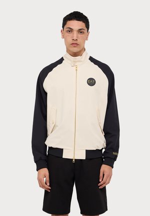 Giacca con zip in crema e blu navy, realizzata in tessuto morbido. Presenta colletto alzato, dettagli neri e logo dorato EA7 sul petto.