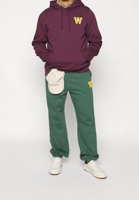 Vínová mikina s kapucí se žlutým logem, doplněná zelenými tepláky s detailem loga. Doplňky zahrnují světle zbarvenou čepici. Celý outfit doplňují bílé tenisky.