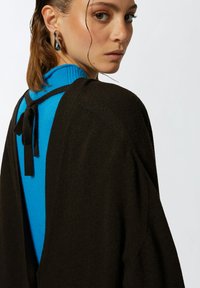 Crni pleteni kardigan s opuštenim krojem, s niskim izrezom na leđima i vezicama, nosi se preko prilagođenog plavog rolaka. Glatka tekstura s kontrastnim bojama.