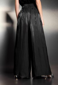 Pantalons larges noirs avec une texture lisse et brillante. Dotés d'une taille élastique froncée, ils tombent jusqu'à la cheville, dévoilant des chaussures à talons.