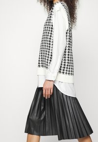 Gilet en pied-de-poule noir et blanc porté sur une chemise blanche boutonnée, associé à une jupe plissée en imitation cuir noir. Détails texturés visibles.