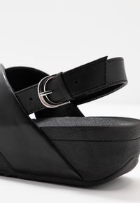 Svart kilklackssandal med texturerad plattformssula och justerbart vristband med en silverfärgad spänne mot en vit bakgrund.