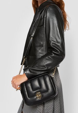 Femme portant une veste en cuir noire et une jupe à motifs, tenant un sac à main noir matelassé avec une bandoulière chaîne dorée et un logo double-B en or.
